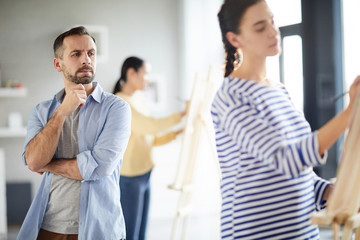 Fototapeta premium Serious teacher looking at easel of one of his students working individually at lesson in studio