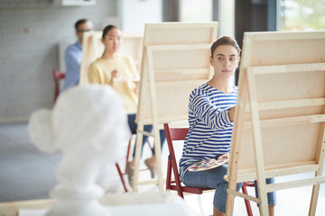 Young female student of painting faculty looking at alabaster human head while sitting in front of easel at lesson
