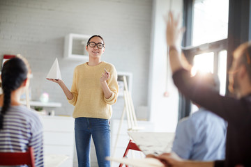 Young casual teacher of painting with paper pyramid asking her students question and pointing at...