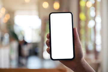 Mockup image of a hand holding and showing black mobile phone with blank white screen in modern cafe