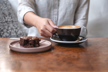 Closeup image of a woman's hand holding and drinking hot latte coffee with brownie cake on the table