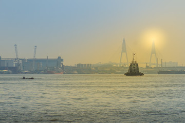 Fototapeta premium Commercial ship sailing on the river near a bridge during sunset at the estuary of Chaophraya river, Samut Prakarn province, Thailand.