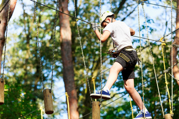 Naklejka na ściany i meble Cheerful cute young boy in white t shirt and white helmet in adventure rope park at sunny summer day. Active lifestyle, sport, holidays for children