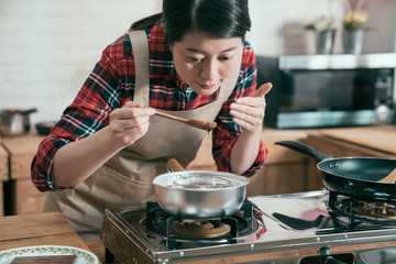 young asian female cook smells the food preparing in the pot while moving it with wooden spoon. beautiful housewife holding melting chocolate in glass bowl on stove in cozy wooden kitchen with love.