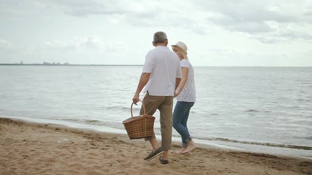 Senior lovers meeting day on beach. Walking and takling on shoreline after picnic. Mature woman in straw hat and with bare feet holding hand of happy retired man.