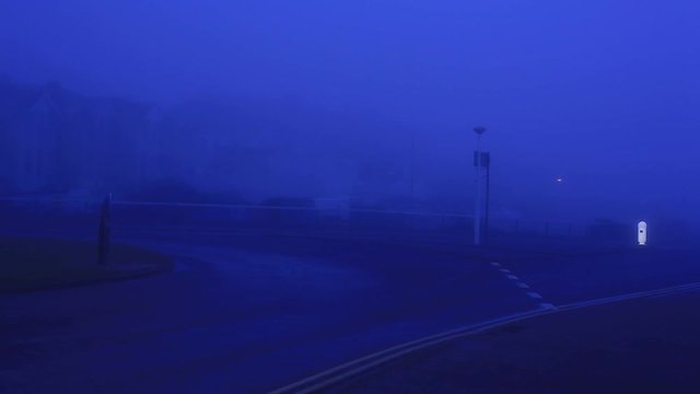 Very foggy morning in UK. Dark blue light. Street lights are still on. Car with headlights still on is driving down an empty road and turns towards the camera, drives past. Footage in HD
