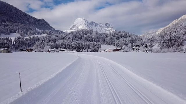 A winter view of a cross country ski trail near Kirchdorf in Tirol, Austria. Wilder Kaiser mountain range in the depths of winter with a deep blanket of soft white snow. Drone accelerating in reverse.