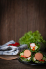 Salad rolls on black plate placed on a wooden table with Fork, White Blue Striped Fabric, lettuce and tomato placed backside.