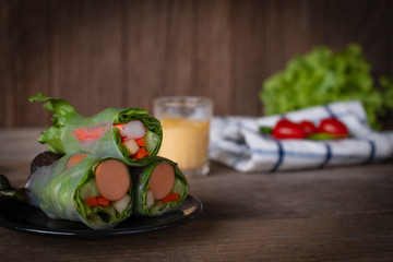 Salad rolls on black plate placed on a wooden table with salad dressing, White Blue Striped Fabric, lettuce and tomato placed backside.