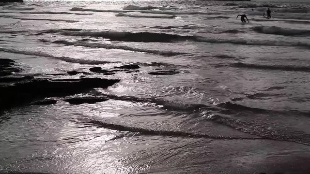Cornish UK coast in summer. Bright sun reflecting glaringly in ocean waters, tide is in. People swimming, boy falls in water, while coming back to a shore; sun will start setting soon. Footage in HD