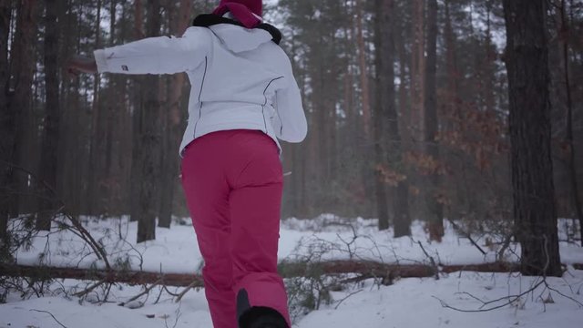 Scared African american young woman dressed warm wearing a red hat, scarf and white jacket running through the snowy forest. Girl running away from the pursuer constantly turns around