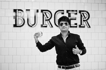 Stylish indian man in sunglasses at fast food cafe with hamburger at hand against burger sign on wall.
