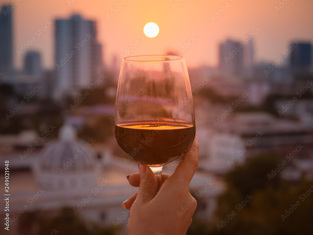 Wall mural happy moment of woman hand with red wine into glasses in a sunset background as on the rooftop at th