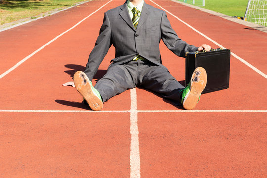 An exhausted businessman in gray suit with green shirt and tie, black briefcase and broken green running shoes sitting on a running track