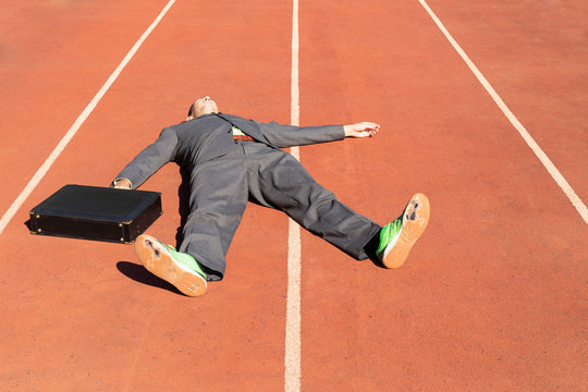 An exhausted businessman in gray suit with green shirt and tie, black briefcase and broken green running shoes lying on a running track
