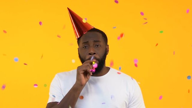 Upset Afro-American man in party hat whistling in horn under falling confetti