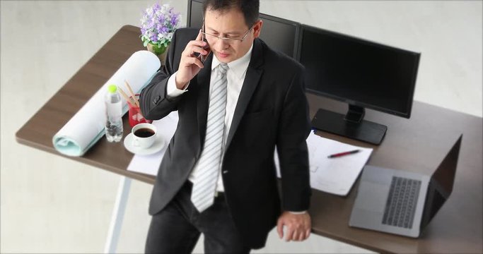  Asian businessman, in black suit, use cellphone, there are notebook, moniter, cofee cup, blueprint on table behind him, he standing in office.