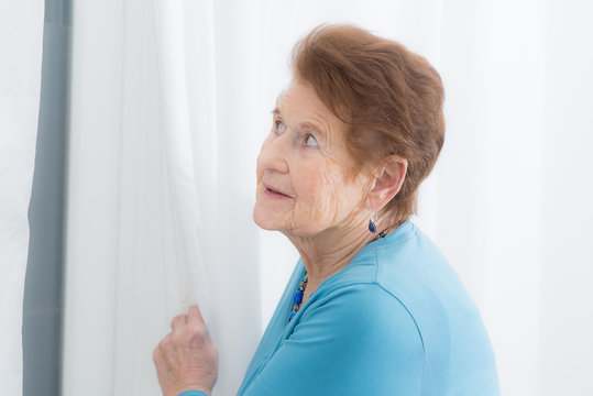 Demente Frau schaut &auml;ngslich hinter dem Vorhang eines Fensters