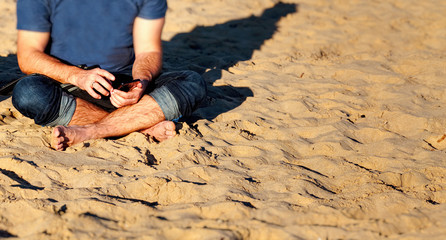 Man sitting on a sand and holding a smartphone in the hands in sunny day. Holiday and summer concept.