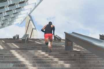 Muscular hispanic Young male dark-skinned athlete running up a flight of stairs with speed, sporty young man in black t-shirt training or working out outdoors while jogging up the steps. Rear view