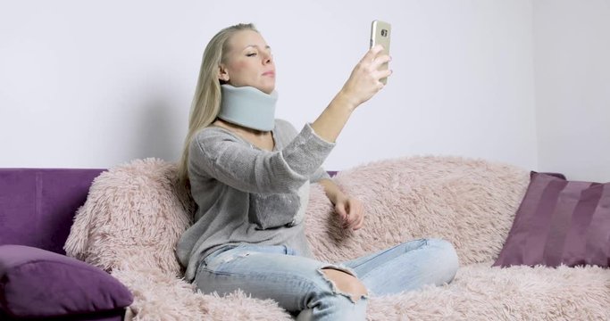 A blonde Caucasian woman at home sitting on her sofa struggles to use her smartphone because of her neck injury and neck brace