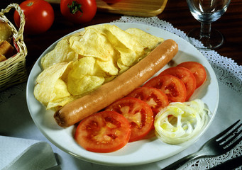Plato con una salchicha frankfurt, patatas chips, y ensalada de tomate y cebolla.