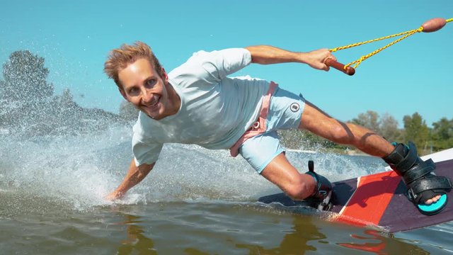 CLOSE UP, TIME REMAP: Handsome Caucasian man wakeboarding does a hand drag and splashes water while speeding past the camera. Smiling wakesurfer riding in the waterski cable park on sunny summer day.