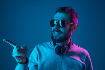 Enjoying his favorite music. Happy young stylish man in sunglasses with headphones listening sound and smiling while standing against blue neon background