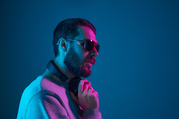 Enjoying his favorite music. Happy young stylish man in sunglasses with headphones listening sound and smiling while standing against blue neon background