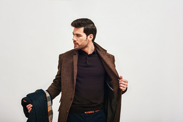 Always perfect. Stylish dark-haired man standing, holding his coat and looking away isolated over...