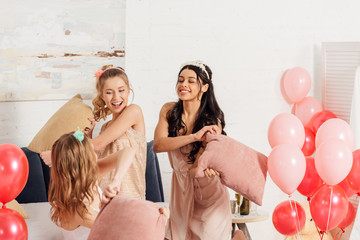 beautiful multiethnic girls in headbands having fun and fighting with pillows during pajama party in bedroom