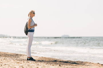 Fototapeta na wymiar Young, beautiful blonde girl walking at the beach. Stylish woman in white pants and sunglasses near sea