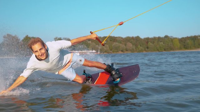 SLOW MOTION, CLOSE UP: Smiling Caucasian wakeboarder sprays the lake water as he speeds past the camera. Cool surfer enjoying a sunny summer day by wakeboarding in a cool park on the tranquil lake.