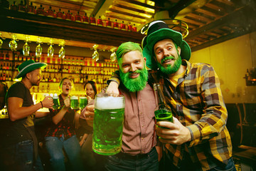 Obraz premium Saint Patrick's Day Party. Happy friends is celebrating and drinking green beer. Young men and women wearing a green hats. Pub Interior.