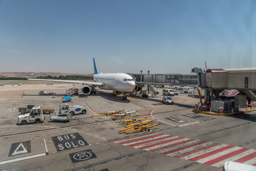 View of the runway of an airport with a plane parked, luggage transport vans and other vehicles