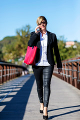 Front view of a elegant business woman holding a red bag walking along the path while using a mobile phone and looking camera outdoors