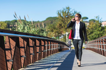 Front view of a elegant business woman holding a red bag walking along the path while using a mobile phone and looking away outdoors
