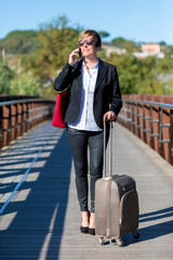 Front view of a smiling elegant business woman wearing red bag and carrying a suitcase on a bridge path while using a mobile phone and looking away in a sunny day