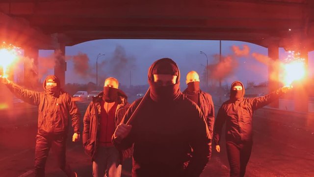 Group of young men in balaclavas with red burning signal flare walking on the road under the bridge, slow motion