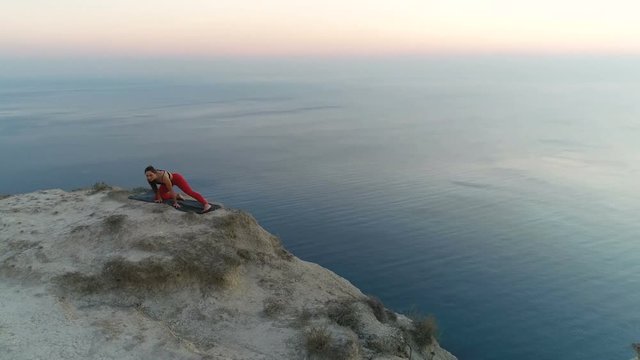 Beautiful young woman is doing yoga stretcing on the top of mountain with sea view at sunset. Aerial footage.