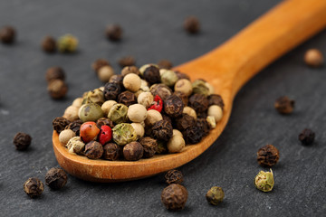 Macro shot of black, green, white and red peppercorn mix in a wooden spoon on a dark stone kitchen board. Shallow depth of field