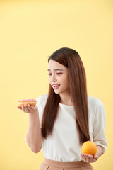 Portrait of a smiling young asian woman choosing between donut and orange isolated over yellow background