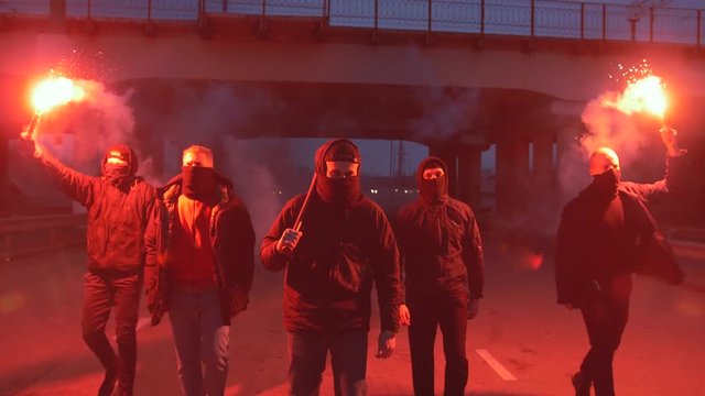 Group of young men in balaclavas with red burning signal flare walking on the road under the bridge, slow motion