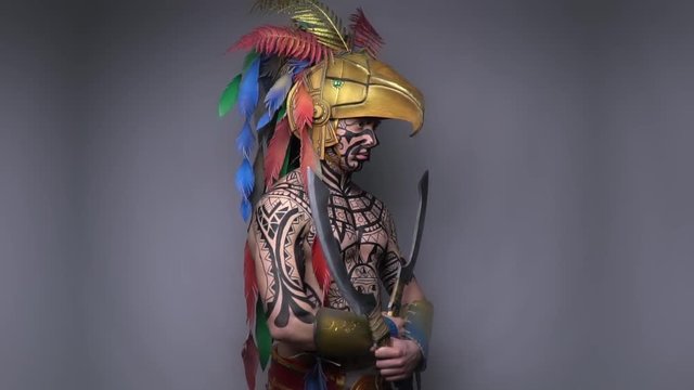 Big buff ancient aztec warrior wearing an eagle helmet with colorful feathers and two blades
