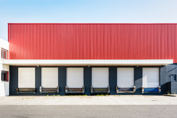 Front view of six truck loading docks at a warehouse in the suburbs of Paris, France, with white roller shutter doors closed under a red corrugated metal siding.