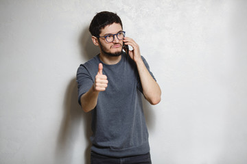 Portrait of young confident man with smartphone in hand showing thumb up, over white textured background.