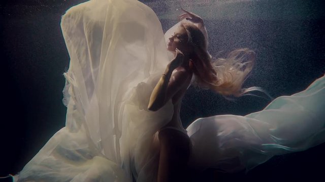 Female slim model is swimming underwater in pool, playing with white cloth