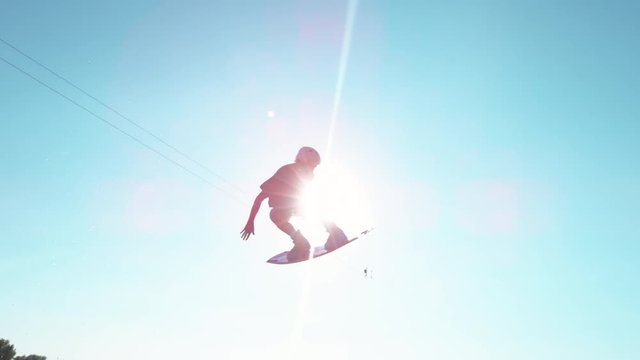 SLOW MOTION, SUN FLARE: Awesome wakesurfer flies across the sunny sky and does a spin trick on a beautiful summer day. Athletic tourist having fun during vacation while wakeboarding on the lake.
