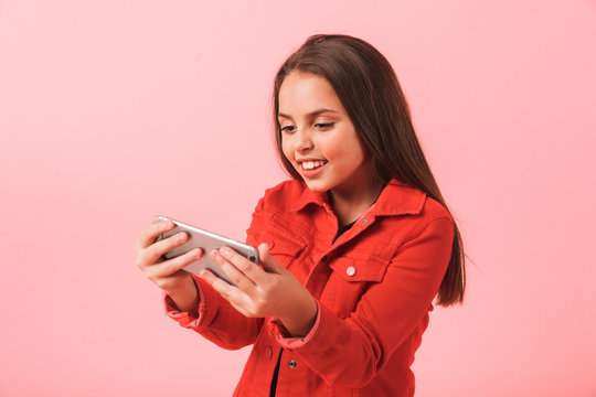 Image Of Happy Little Girl In Casual Playing Video Games On Cell Phone While Standing, Isolated Over Red Background