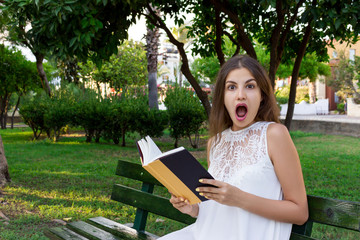 Surprised young girl with a wide opened mouth is reading a book in the park